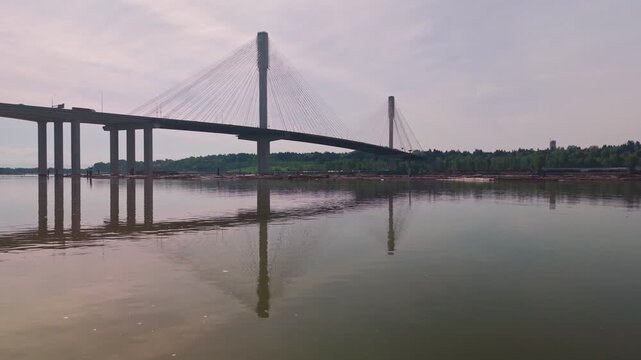 Cable-stayed bridge towers dominate the horizon as camera glides over gravel shoreline and glassy Fraser River water toward massive architectural span approaching