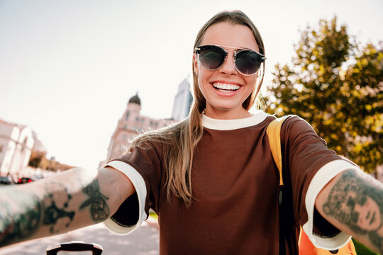 Young woman taking a joyful city selfie with sunglasses and tattoos while traveling and smiling on a sunny day