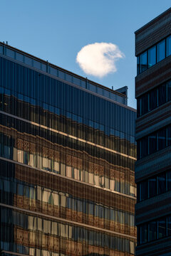 glass office facade rises between corporate buildings under blue sky as reflected windows frame a small cloud above urban architecture
