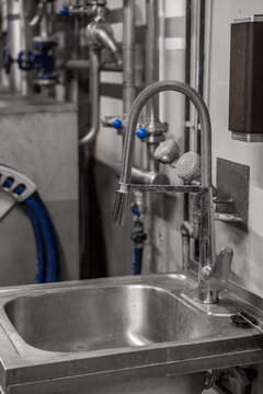 Industrial sink with flexible faucet and sprayer in a brewery production area, surrounded by stainless steel pipes and equipment, with copy space
