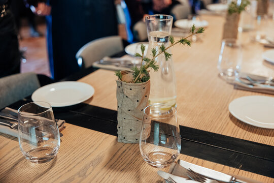 Wooden restaurant table with plates, glasses, cutlery, and small evergreen centerpiece in warm ambient light, with copy space