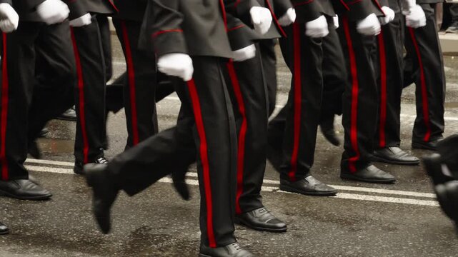 Close-up of uniformed marchers stepping in unison on a wet asphalt street, black trousers with red side stripes and polished shoes in disciplined formation during parade