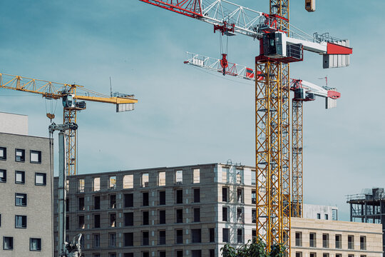 multiple construction cranes watch over a city building site, with urban development and architecture held beneath a soft gray sky