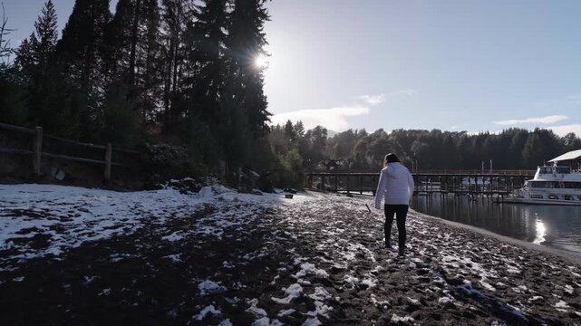 A woman in a white jacket walks along a snow-dusted beach beside a marina in Bahia Mansa. The sun shines through the trees, creating a serene winter setting. Suitable for travel and lifestyle content.