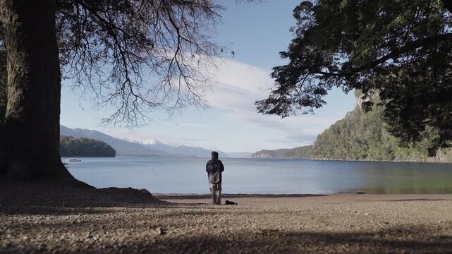 A lone man stands by the calm waters of Lago Nahuel Huapi, framed by trees. Ideal for themes of solitude and nature exploration.