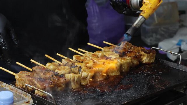 A street food vendor using a gas torch to grill delicious Asian dim sum or siomay skewers on a hot flat griddle.