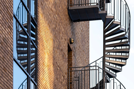 metal spiral stairs rise beside brick facade and balconies, where urban architecture and shadowed exterior details frame a narrow building passage