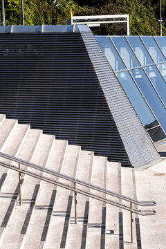 concrete stairs and metal handrail lead beside black facade and skylight, where urban architecture creates sharp shadows across an exterior entrance