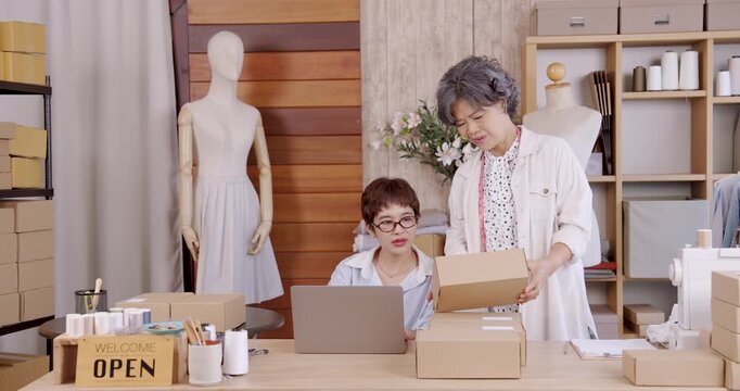 Senior asian woman showing parcel to daughter while discussing order processing inside fashion boutique studio small family business teamwork ecommerce entrepreneurship online shop packaging shipping