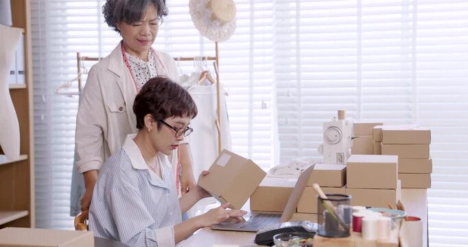 Asian senior female mother standing beside daughter sitting at table checking garment boxes while preparing shipment inside boutique atelier family fashion small business teamwork ecommerce process
