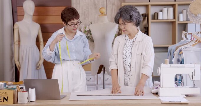 Senior asian mother and adult daughter measuring fabric with tape on worktable near sewing machine, teamwork creating handmade garment pattern for small family boutique fashion business online shop