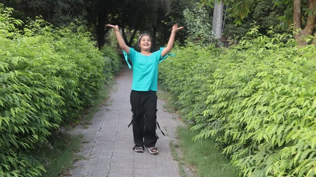 Happy Indian girl walking with raised hands on a peaceful green pathway during summer outing. Cheerful outdoor lifestyle moment surrounded by fresh plants and natural scenery.