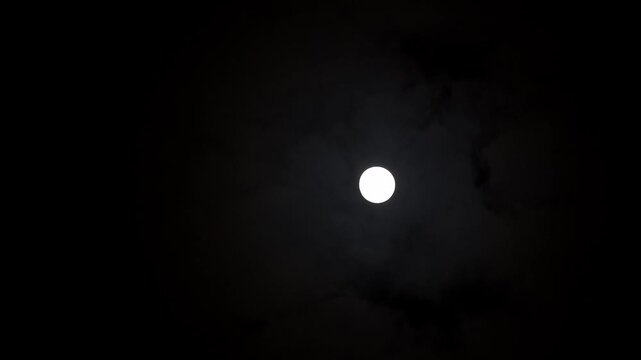 A serene, high-contrast night sky shot featuring a bright full moon surrounded by dark, wispy clouds slowly drifting by, creating a mysterious and tranquil atmosphere.