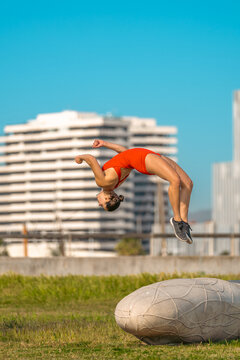Side view of an athletic young woman wearing a red jumpsuit mid-air performing a back flip or back handspring in an urban park with city buildings