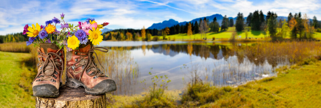 Ein Paar Wanderstiefel gef&uuml;llt mit einem Strau&szlig; Blumen, steht auf einer Blumenwiese vor einem malerischen See im Allg&auml;u - FOTOMONTAGE