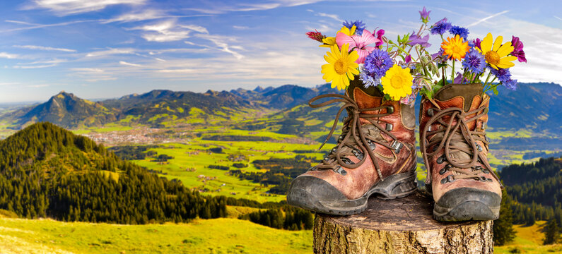 Ein Paar Wanderstiefel gef&uuml;llt mit einem Strau&szlig; Blumen, steht auf einer Wiese vor dem Bergpanorama im Allg&auml;u - FOTOMONTAGE