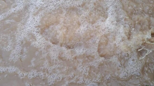 ​Dirty muddy water flowing and bubbling with white foam, close up high angle shot.