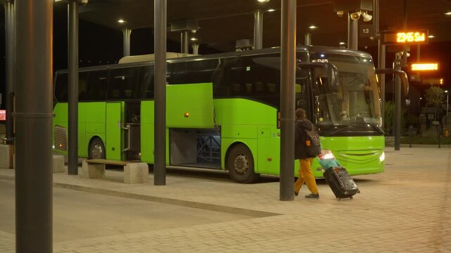 Tourist passenger with rolling suitcase and backpack walking to intercity bus at night station terminal. Traveler preparing for public transport trip journey, commute or holiday vacation.