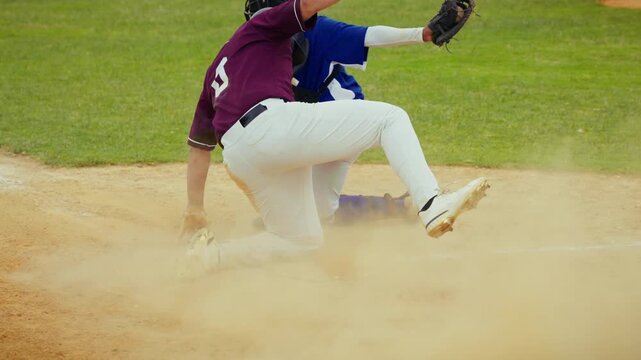 A determined baseball player runs and executes a perfect slide into home plate, kicking up a cloud of dust. The catcher watches the dynamic play unfold on the field.