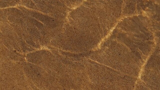 Clear shallow water flows over golden sand, creating dynamic rippling light patterns and caustic shadows on the seabed floor with a serene and meditative atmosphere.