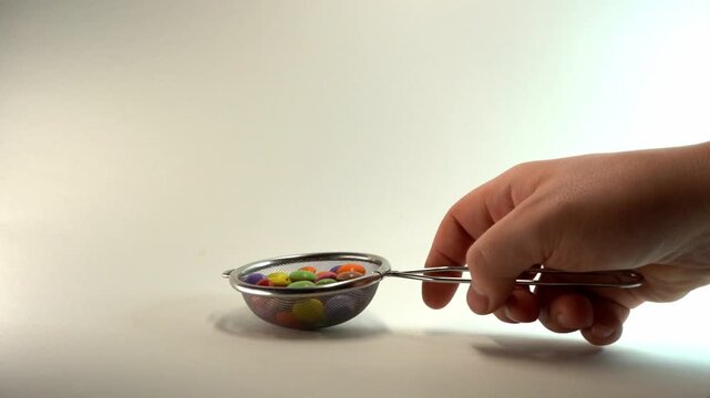 Close-up of a hand using a metal mesh strainer to scoop and manipulate colorful candy-coated chocolate drops on a clean white background, perfect for culinary and food-related concepts.
