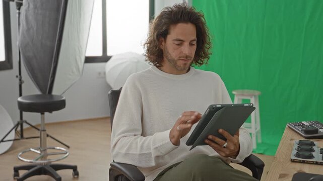 Man photographer taps tablet with hands in studio green screen and softbox visible; focused creativity.