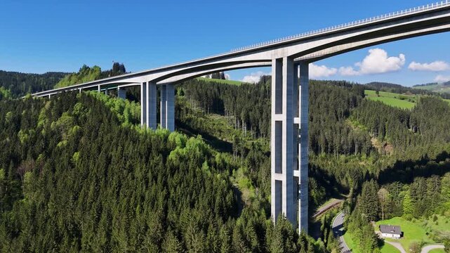 Aerial view of the Lavanttal Bridge with its massive concrete pillars spanning across a lush green forested valley under a clear blue sky in Wolfsberg, Carinthia, Austria.
