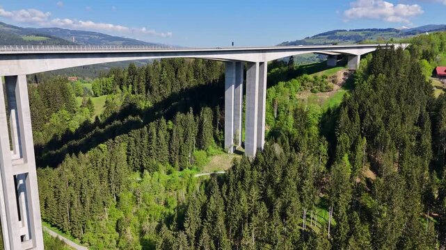 Aerial view of the Lavanttal Bridge spanning across a deep valley with dense pine forests and rolling green hills under a blue sky in Wolfsberg, Carinthia, Austria.