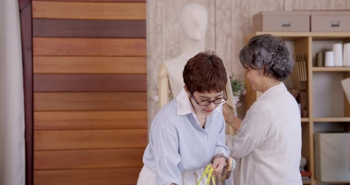 Senior asian mother and adult daughter pinning skirt fabric on mannequin waist, teamwork developing handmade garment design for family boutique fashion studio with textile pattern and sewing tools