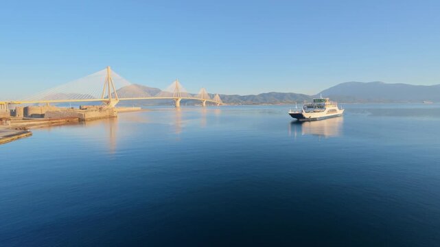 Wide angle establishing shot of a ro-ro ferry sailing through calm harbor waters with mountains and a massive bridge in the background in Greece, Europe.