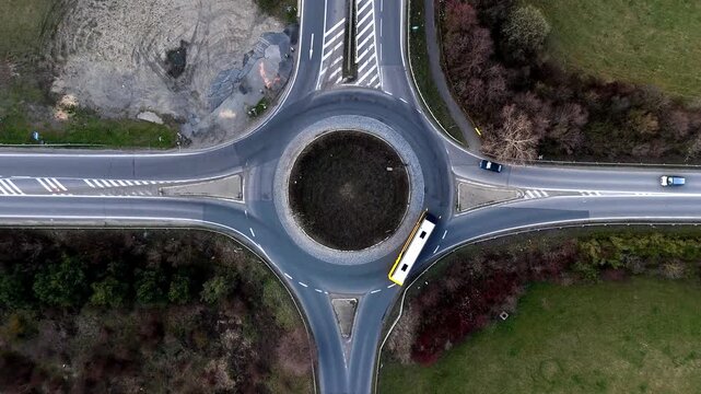 Aerial drone view from directly above of a public transport bus driving through a circular road intersection (roundabout) in a rural or suburban landscape.