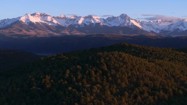 Aerial drone shot tracking left to right across snow-capped San Juan Mountains and Wilson Peak, Colorado. Blue hour alpenglow on peaks with forested foreground providing depth and parallax.