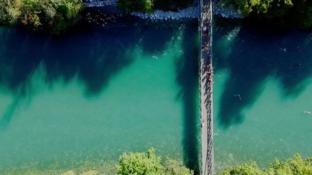 Cinematic vertical drone shot rising slowly over Schonausteg bridge in Bern. People jumping into the turquoise Aare river seen from a top down birds-eye-view during a summer evening.
