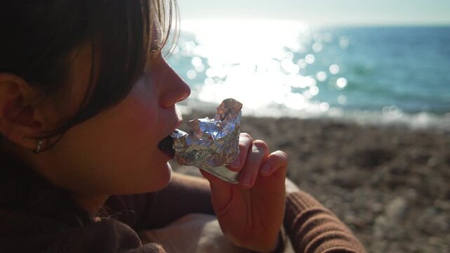 Woman eating beach chocolate as she relaxes by the sea under the bright sun on a peaceful day