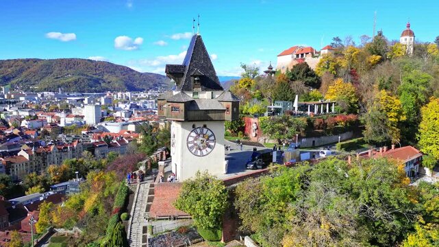 Aerial view of the Graz Clock Tower on Schlossberg hill surrounded by autumn foliage and historic city rooftops under a blue sky, Graz, Styria, Austria.