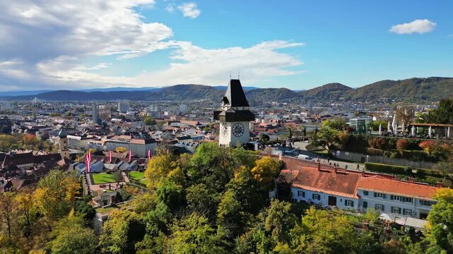 Aerial view of the historic Graz Clock Tower on Schlossberg hill surrounded by autumn foliage and the urban skyline under a cloudy sky in Graz, Styria, Austria.