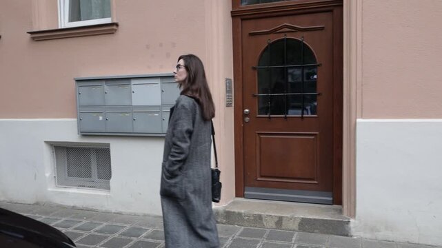 Woman in stylish grey coat walks past apartment building with mailboxes and wooden door