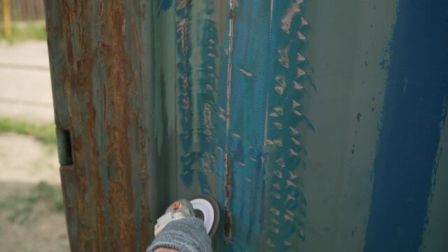 Worker's hand in a glove uses an angle grinder with a flap disc to remove old blue paint and rust from a corrugated metal surface. Close up of metal restoration and surface preparation for painting.