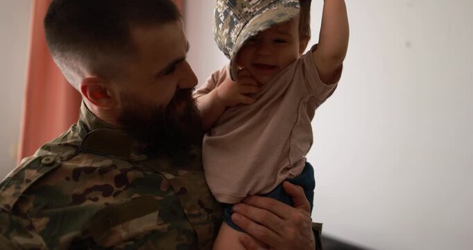 American soldier man in military uniform hugs his little son at home while baby boy playing with his hat - Emotional and happy homecoming concept