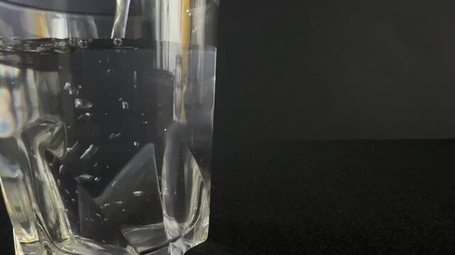 Clear glass filled with water shows continuous pouring action as liquid cascades into the glass, creating ripples and bubbles against a dark background