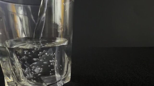 Clear glass tumbler filled with water as liquid is poured in, showcasing the flow and interaction with ice cubes against a dark background