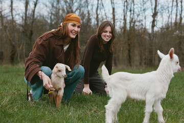 Fototapeta premium Two girls share a playful moment with a kid goat outdoors in the countryside, capturing a lifestyle scene with friends, animals and nature on a grassy field, bright smiles and candid interaction