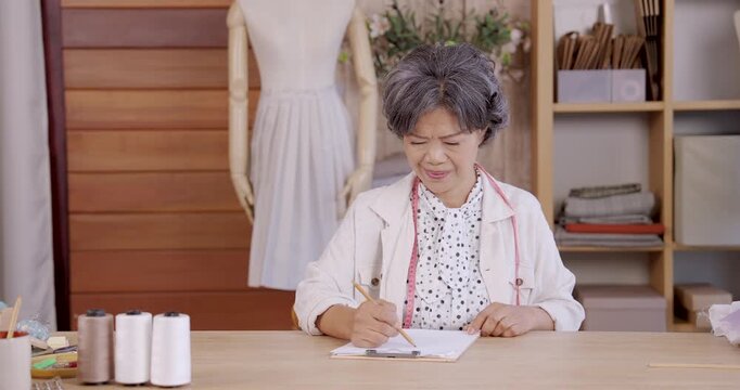 Senior asian woman concentrating while writing notebook at desk with sewing tools and mannequin in studio, managing handmade fashion tasks for family boutique business and preparing online shop orders