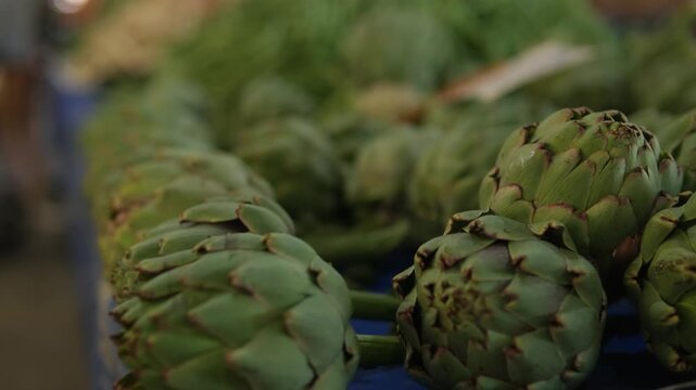 Fresh artichokes on market stall, closeup row of green heads in wooden crate, soft warm light, blurred shoppers and produce in background, rustic organic
