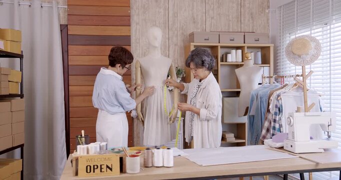 Adult asian daughter adjusting skirt fabric on mannequin while senior mother supporting, teamwork designing handmade fashion garment for family boutique business with sewing machine and online shop