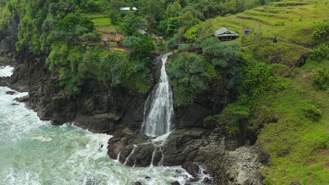 Aerial view of Banyutibo Water Fall cascading from a rocky cliff into the ocean with lush green hills and small huts in Girisubo, Special Region of Yogyakarta, Indonesia.