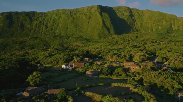 Aerial drone high-angle view of a traditional village with stone houses and terracotta roofs, nestled in a lush green valley at the base of dramatic volcanic mountain slopes, Flores Island, Azores.