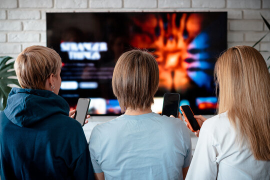 A group of three friends gathered in a cozy living room, watching a movie on a large TV while engaged with their smartphones. The scene captures modern entertainment habits.