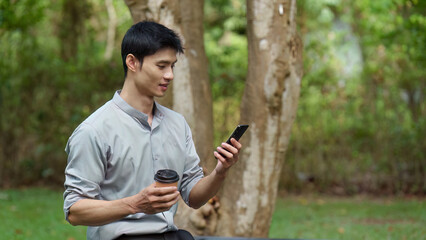 Fototapeta premium East Asian man working remotely on smartphone with coffee in park