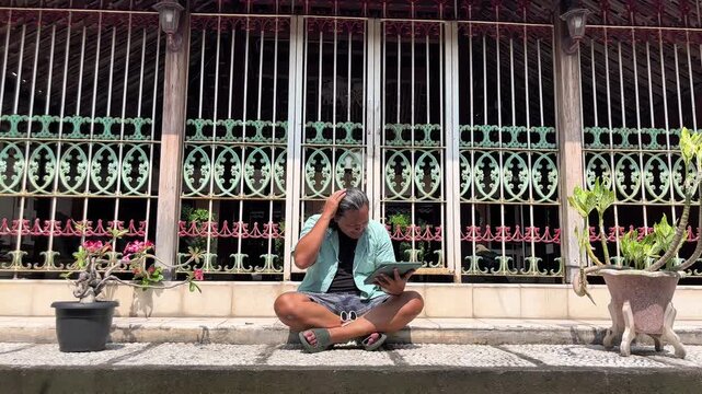 Adult asian man is sitting in front of heritage house in Kotagede while watching online worldcup championship by using an ipad